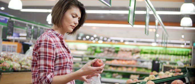 Brown haired woman in fruit and veg aisle of supermarket looking quizically at an apple in her hand. 