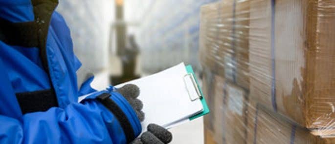 Close up of hands checking paperwork on a clipboard in front of wrapped packages inside a warehouse full of containers.