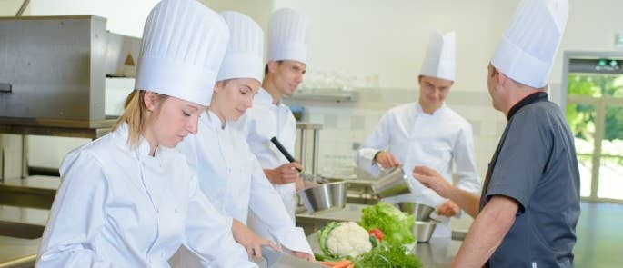 Group of young chefs gather round workstation while chef master explains cooking techniques to them