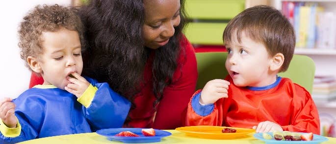 Female nursery teacher helping two children have fruit snacks.