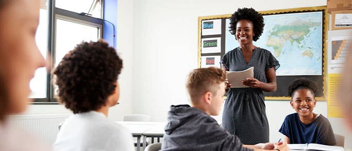 teacher stood at the head of a classroom smiling whilst handing out papers